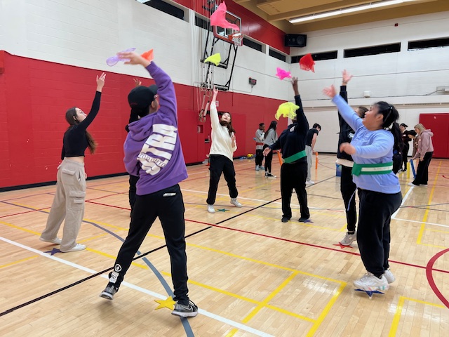 A small group of kinesiology and high school students are each holding and tossing a colourful scarf in the air.  Each person moves to the right while the scarves are in the air and attempts to receive the new scarf before it falls to the ground, challenging their hand-eye co-ordination and movement skills.