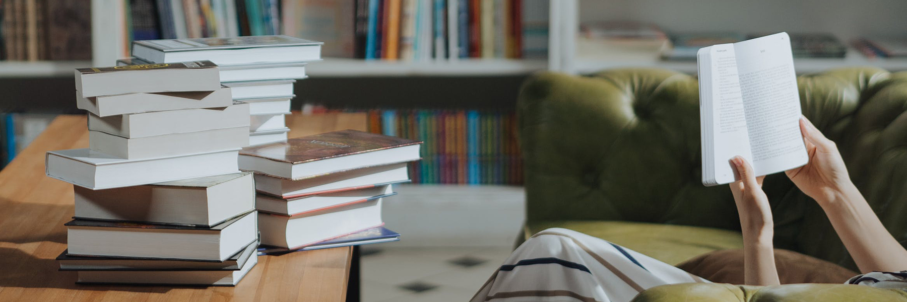person reading a book on couch with pile of books nearby BANNER